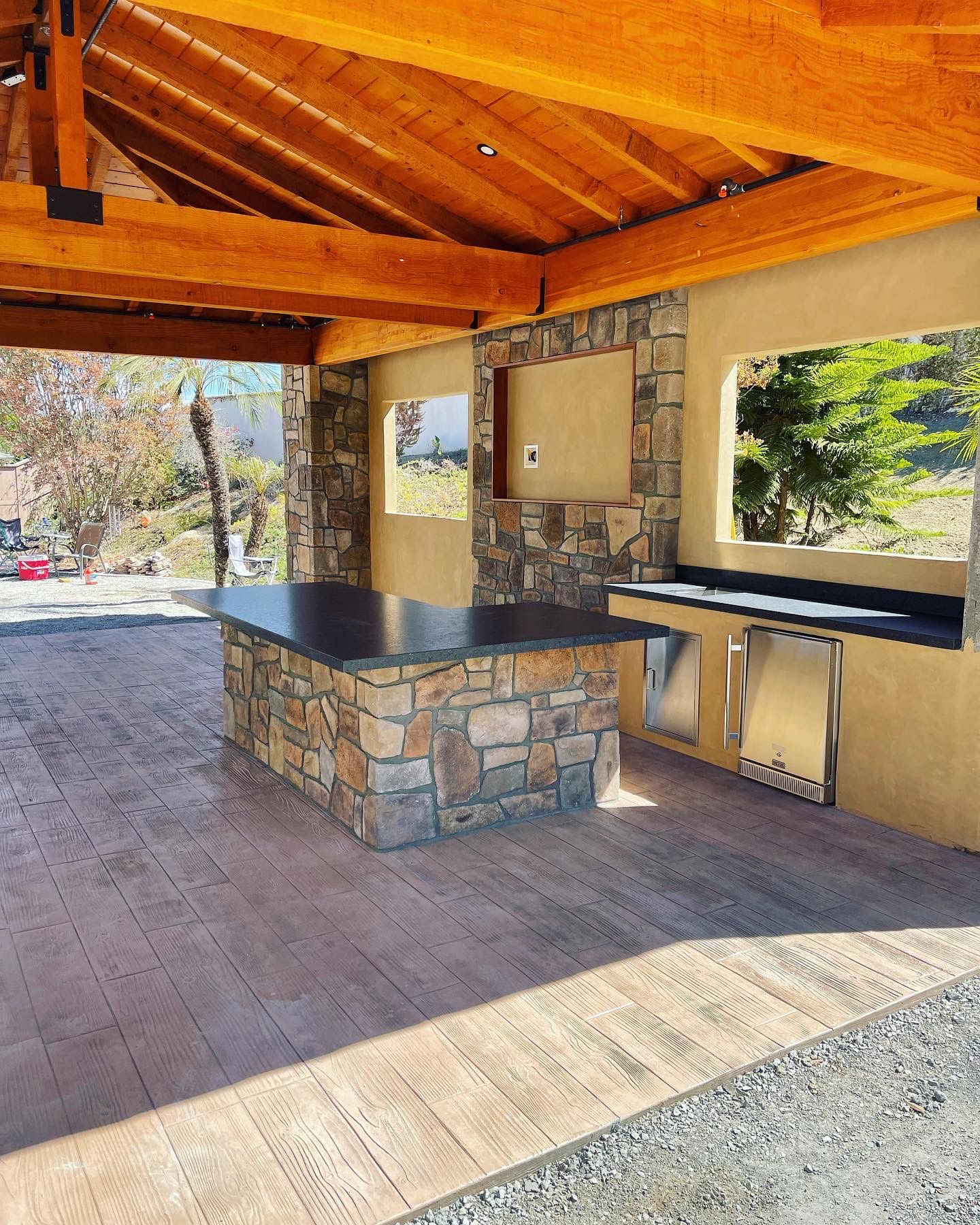 Outdoor stone barbecue pavilion with wooden ceiling, countertop, and sink under open shade