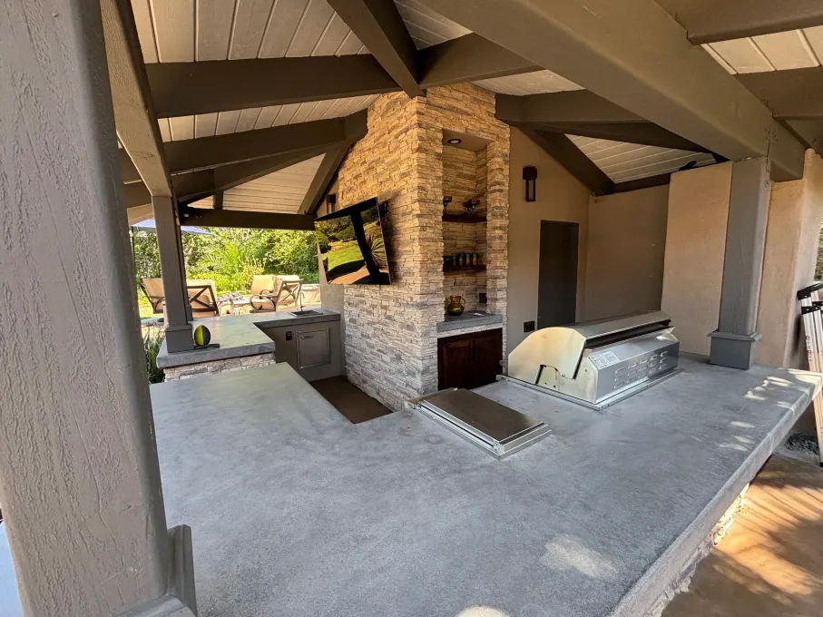 Covered outdoor patio with stone fireplace, white seating, and a ceiling fan under a sloped roof.