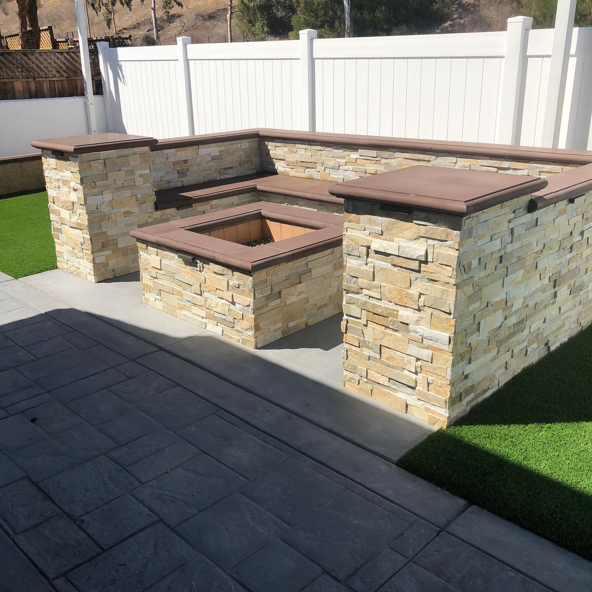 Stone outdoor seating and fire pit on a patio beside a white fence and lawn.