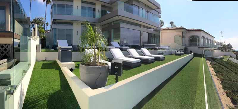 Modern home patio with lounge chairs, potted plants, and artificial grass beside a pool or waterway
