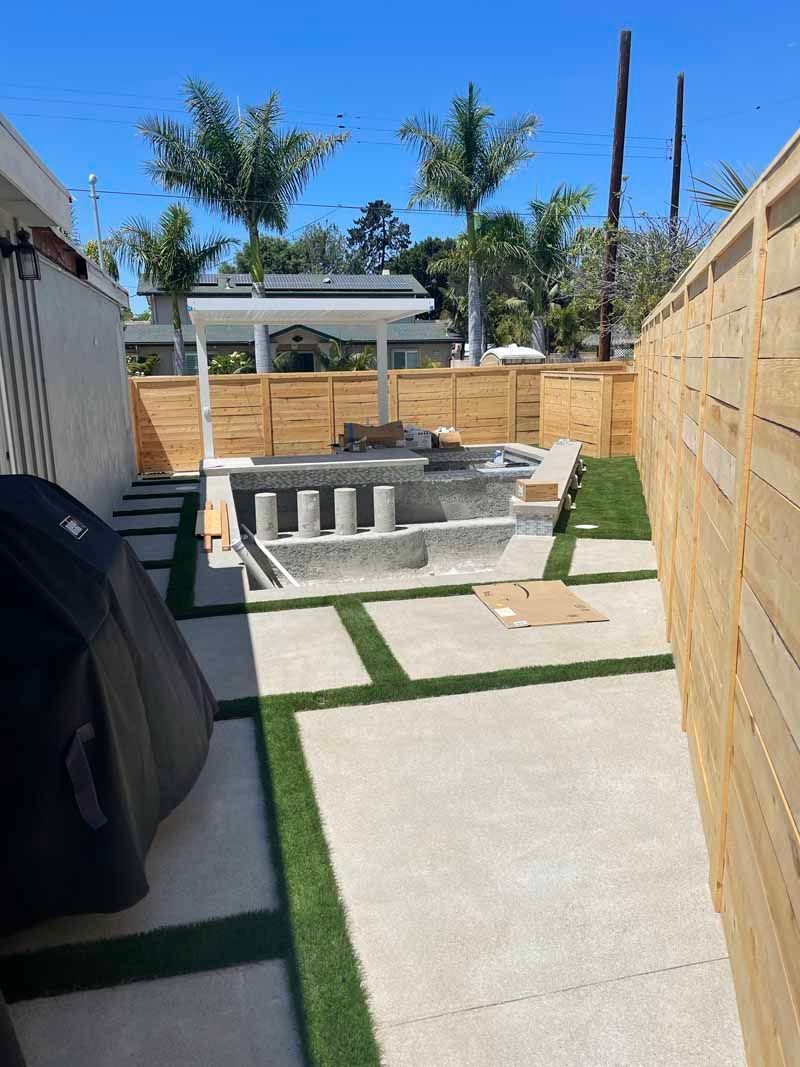 Backyard patio with a concrete path, pool, and wooden fencing under a clear blue sky.