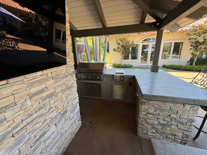 Covered outdoor kitchen with stone counters, built-in grill, and a backyard house in the background