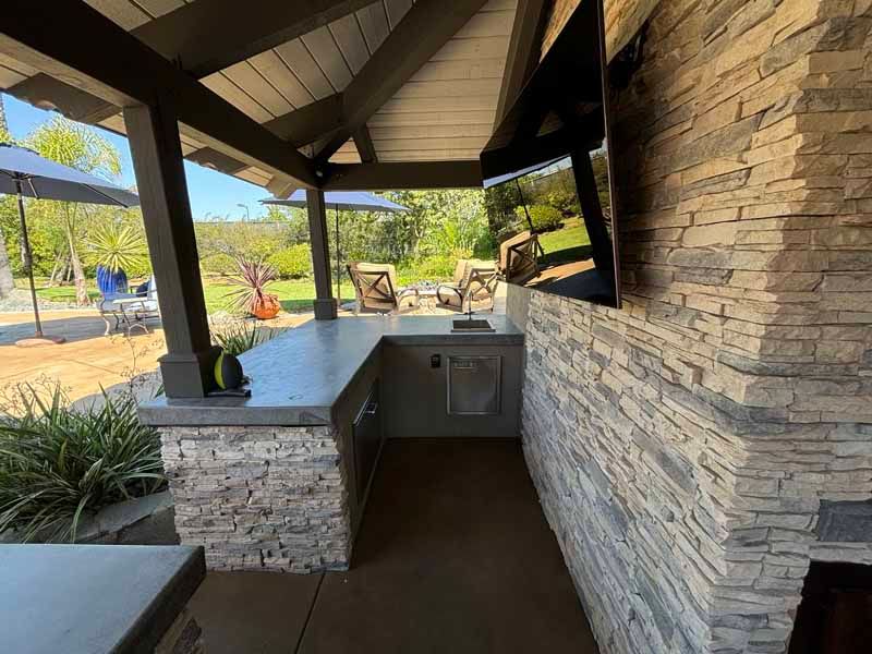 Covered outdoor patio with stone walls, built-in grill, and mounted TV overlooking a landscaped yard.
