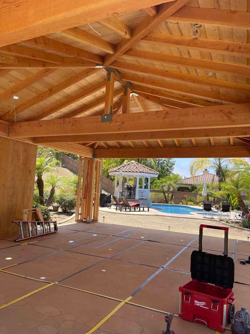 Open-air wooden pavilion with a red rolling suitcase on the floor, overlooking a sunny courtyard and pool.