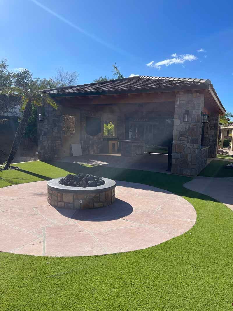 Stone fire pit on a circular patio beside a rustic building, with green lawn under a sunny blue sky