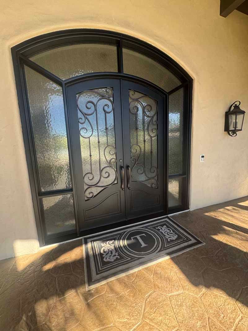 Front entrance with ornate black double doors, arched glass window, and a welcome mat on a sunlit porch