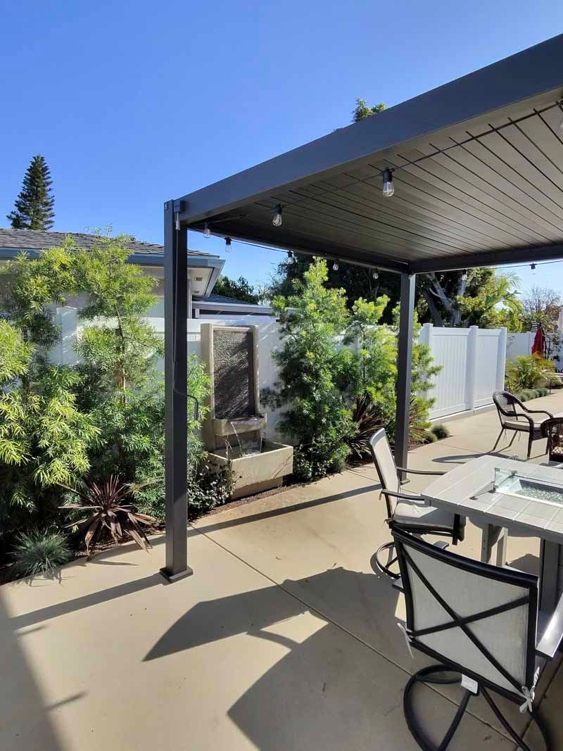 Patio with pergola, table and chairs, surrounded by lush greenery under a clear blue sky.