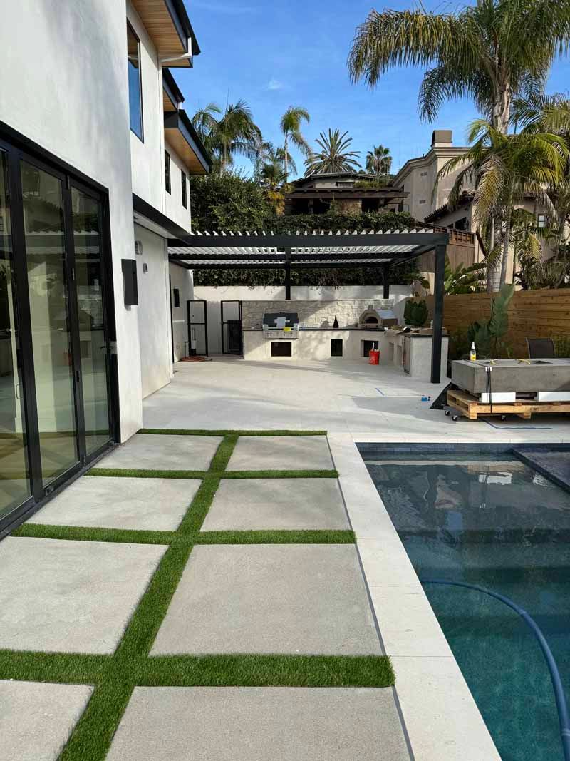 Modern patio with pool, white pavers, outdoor kitchen, and lounge seating beside a house