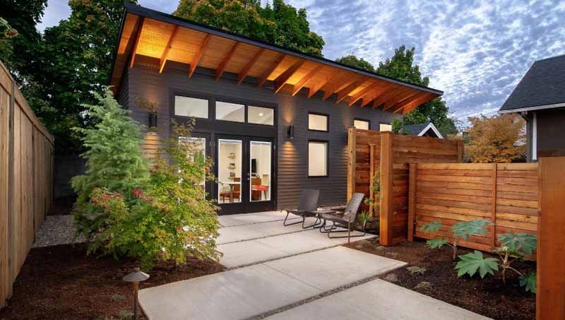 Modern backyard patio with a small gray house, slanted wood roof, pavers, plants, and lounge chairs.