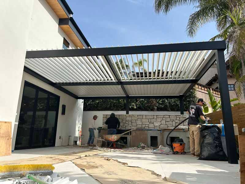 Covered patio with two people building a grill area beside a white house
