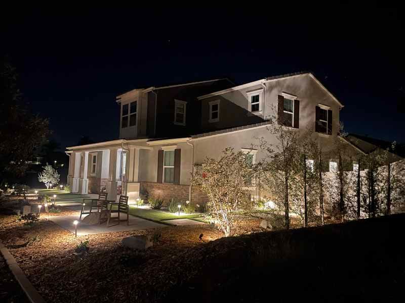 Nighttime view of a lit two-story house with porch lights and bushes in the yard