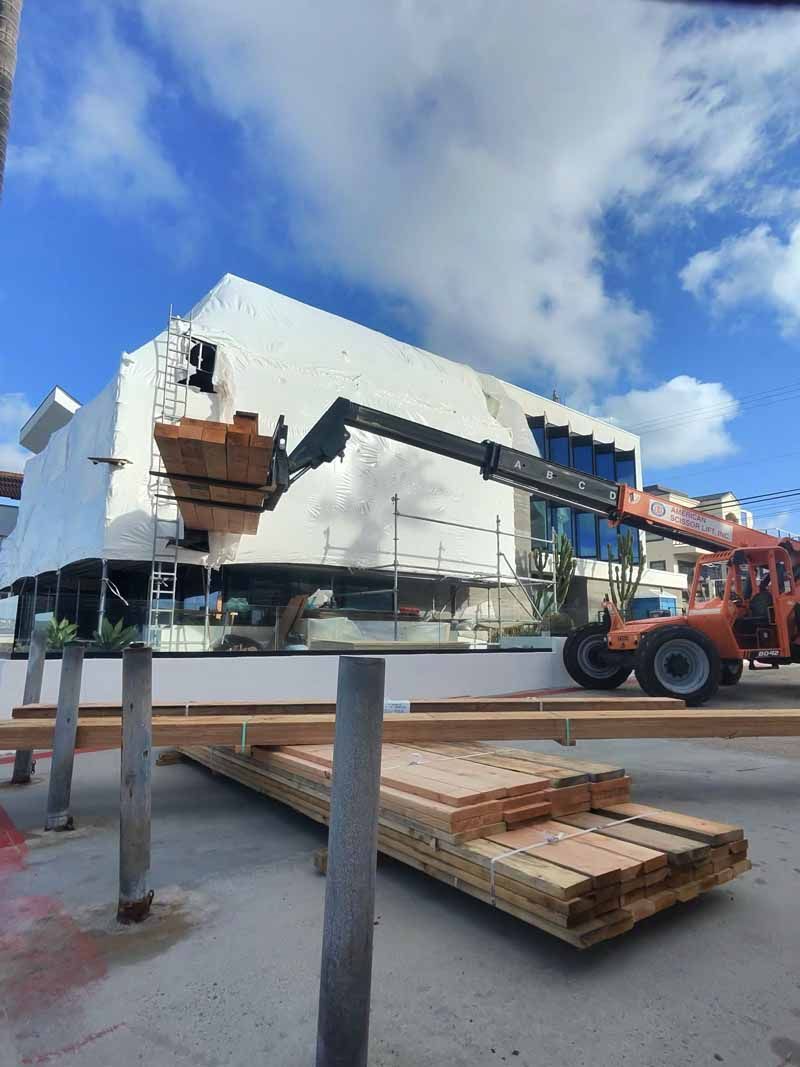 Construction lift moving bundled lumber near a white building under a cloudy sky