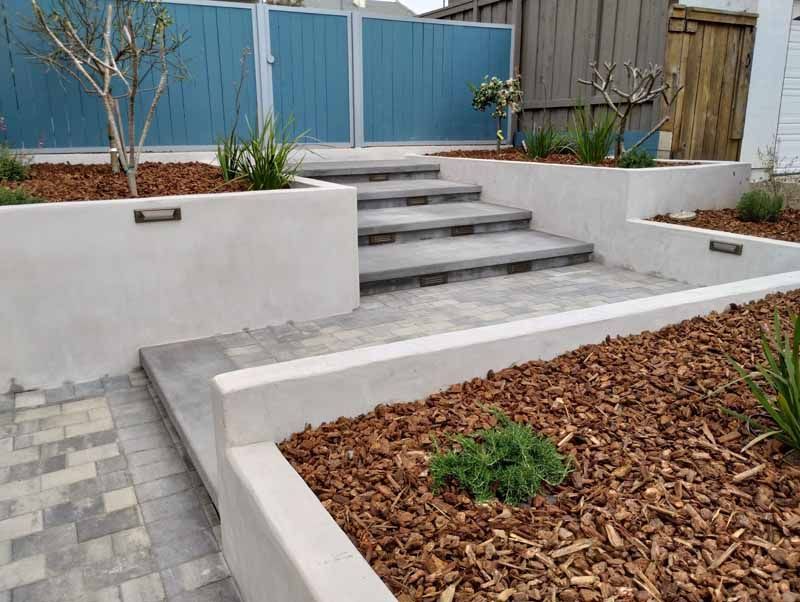 Modern front yard with concrete steps, white retaining walls, blue fence, and wood-chip planter beds
