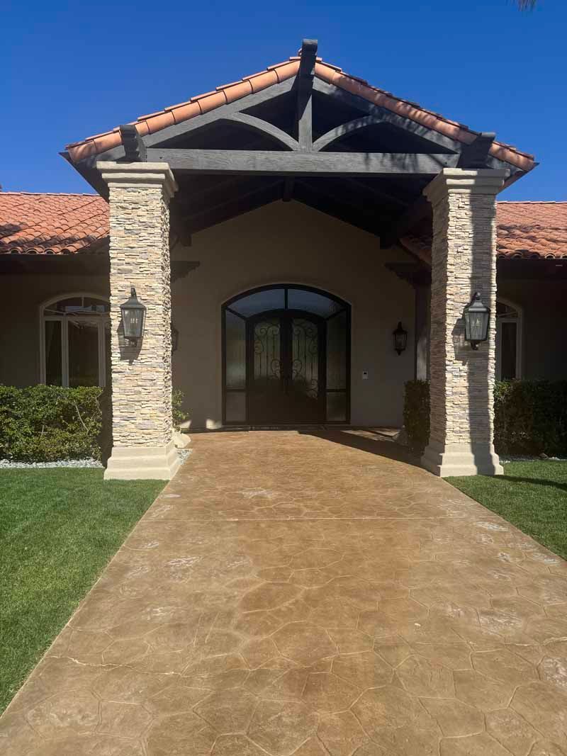 Front entrance of a stucco house with stone columns, arched doorway, and a long tan walkway.