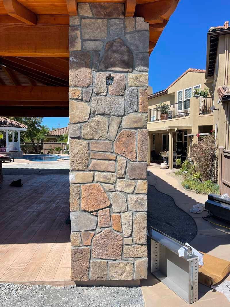 Stone column under a wooden roof beside a sunny courtyard and walkway with buildings in the background