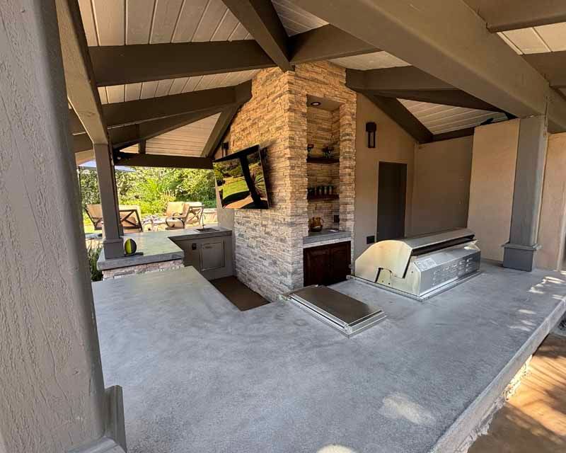 Covered outdoor patio with stone fireplace, built-in grill, and beige columns under a wooden roof.