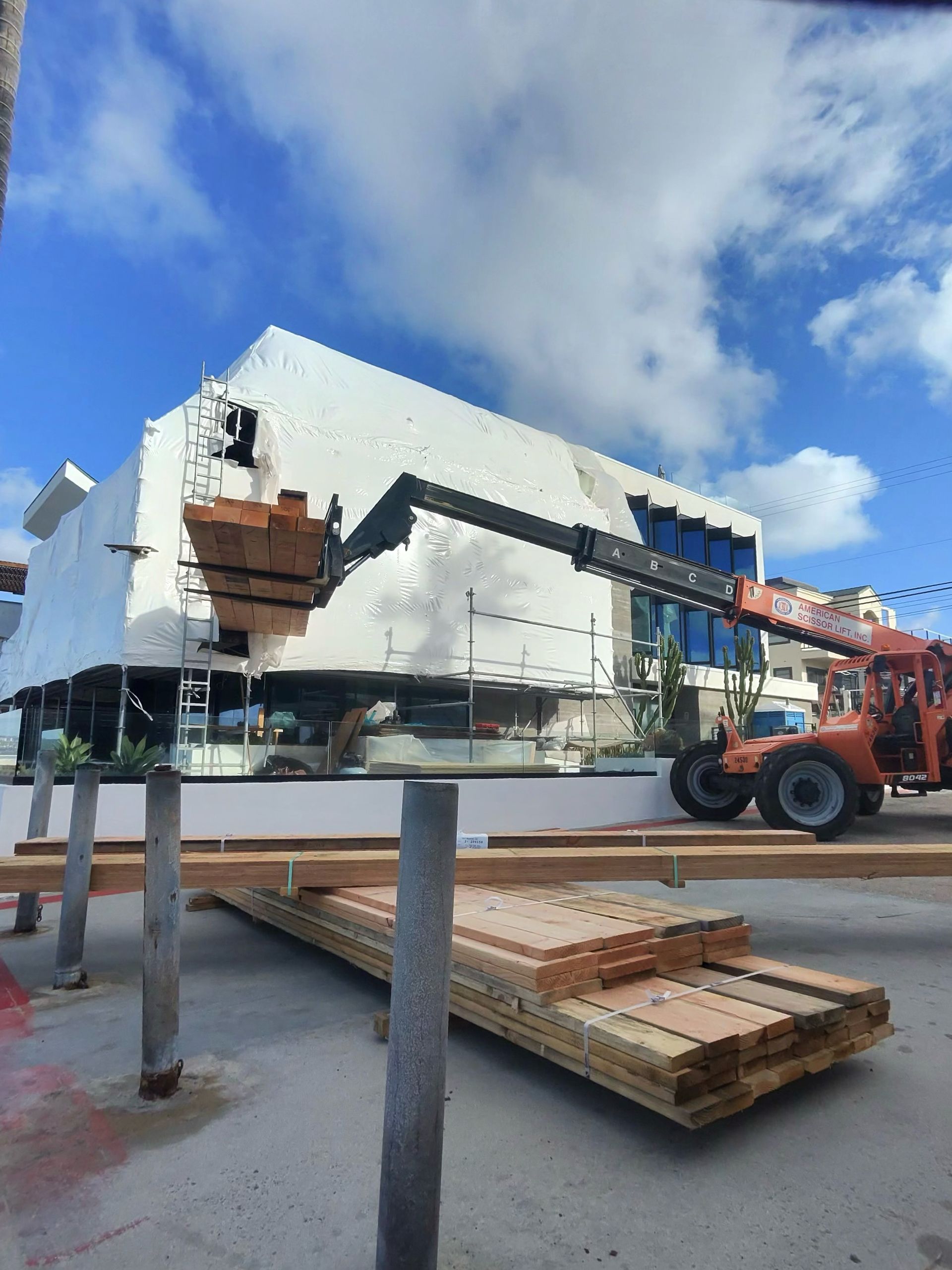 Construction site with a white building, orange lift, and stacked lumber under a blue sky.