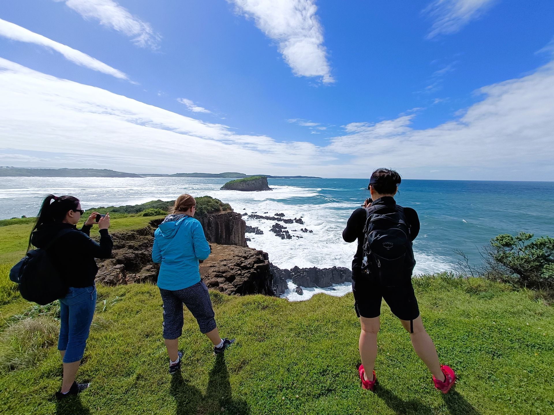 Three people are standing on top of a grassy hill overlooking the ocean.