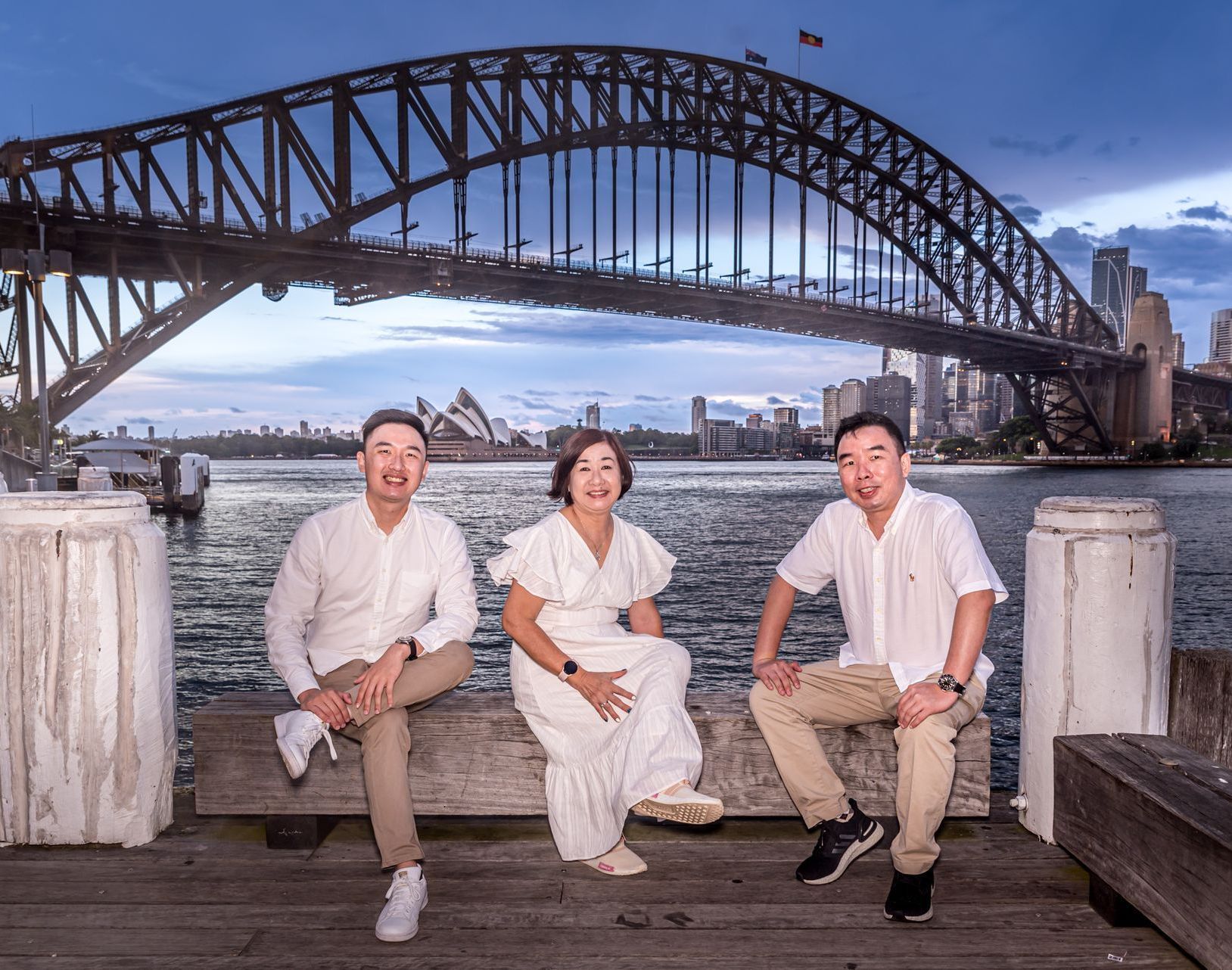 Three people are sitting on a bench in front of a harbor bridge.