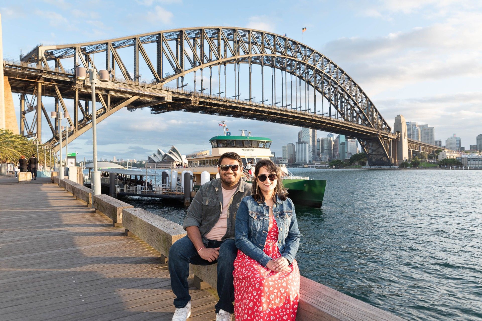 A man and a woman are sitting on a pier in front of a bridge.