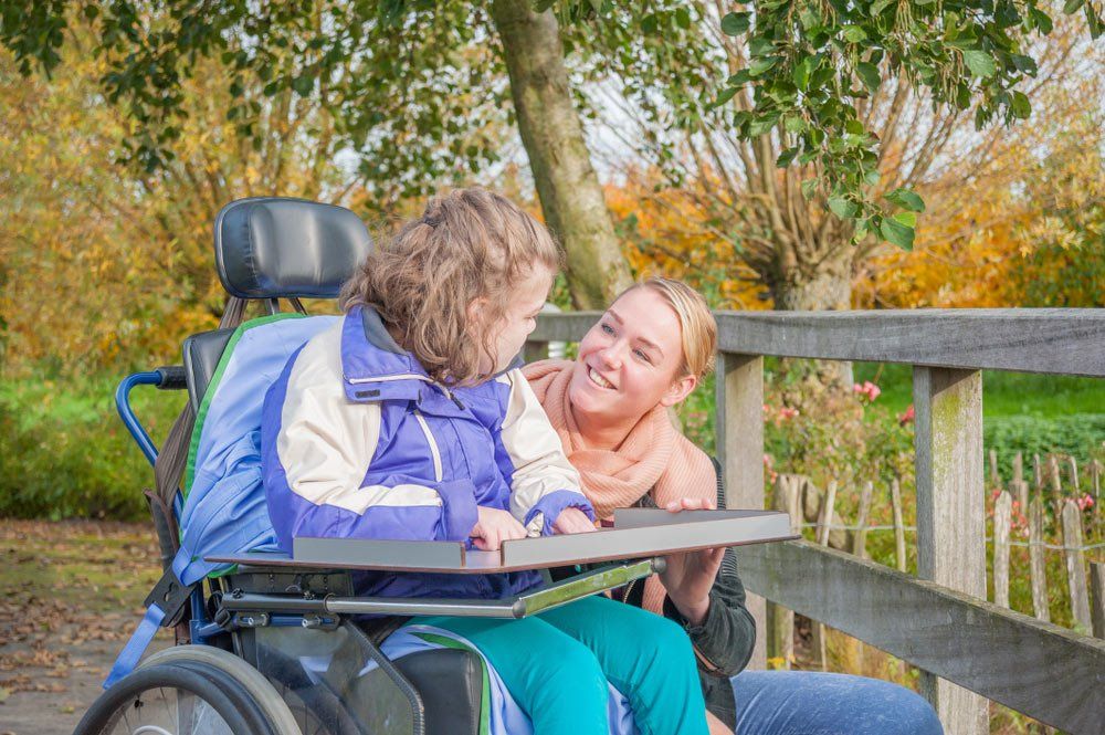 A Disabled Child In A  Wheelchair Relaxing Outside Together With Care Worker — iCan Support Services Kawana QLD