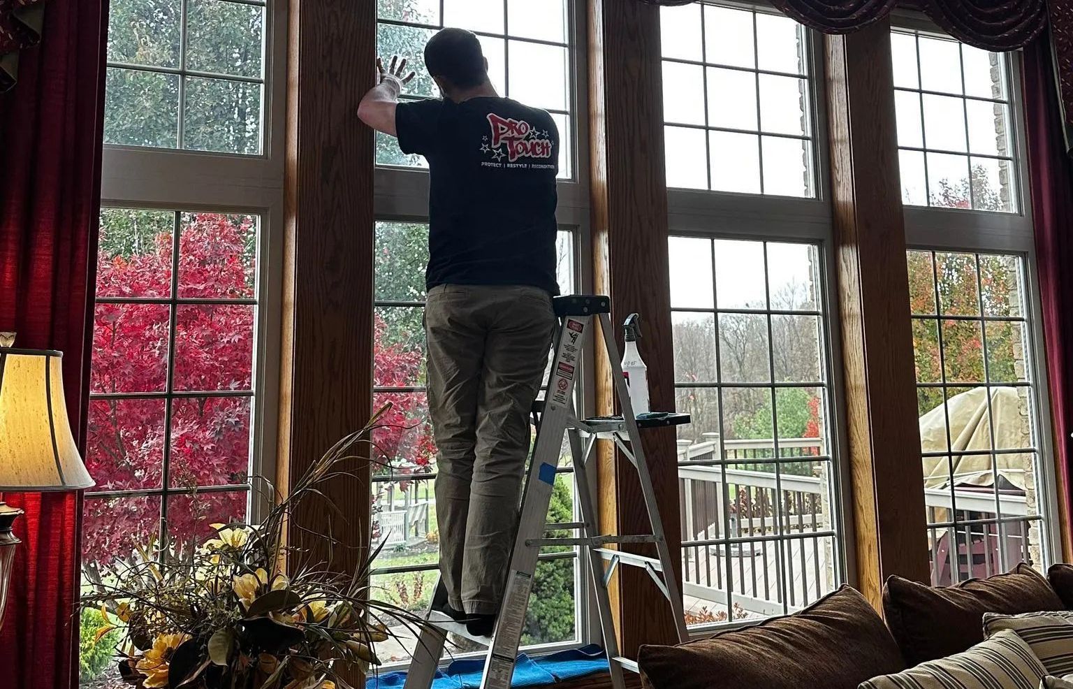 Person on ladder installing window tint in a room with large windows, red curtains, and outdoor view.