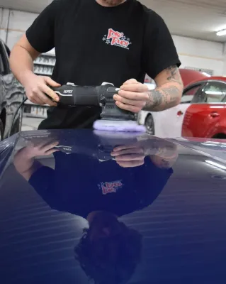 Person polishes a blue car roof with an electric buffer in a garage.