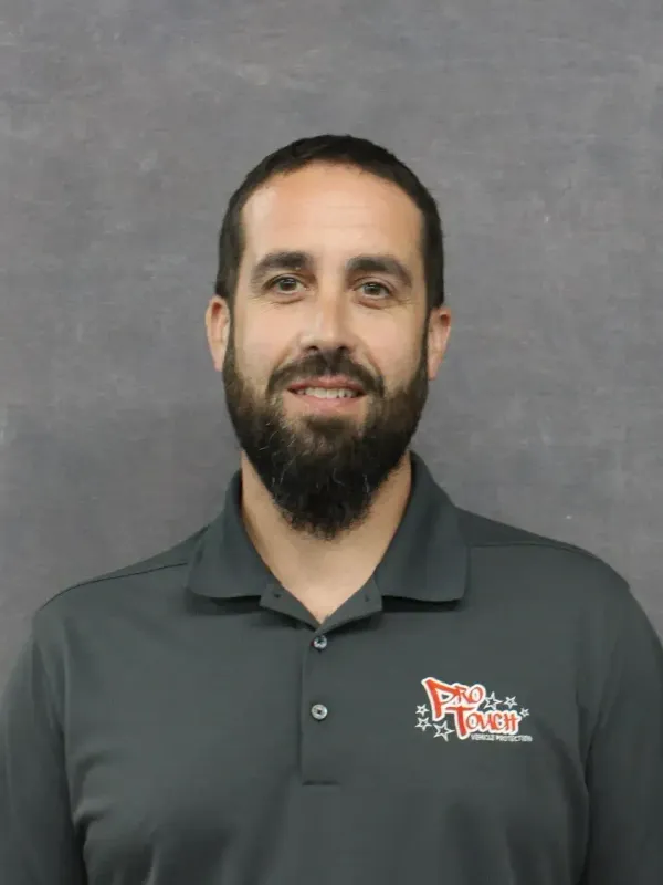 Man with beard wearing gray polo shirt smiling, logo on shirt. Gray background.