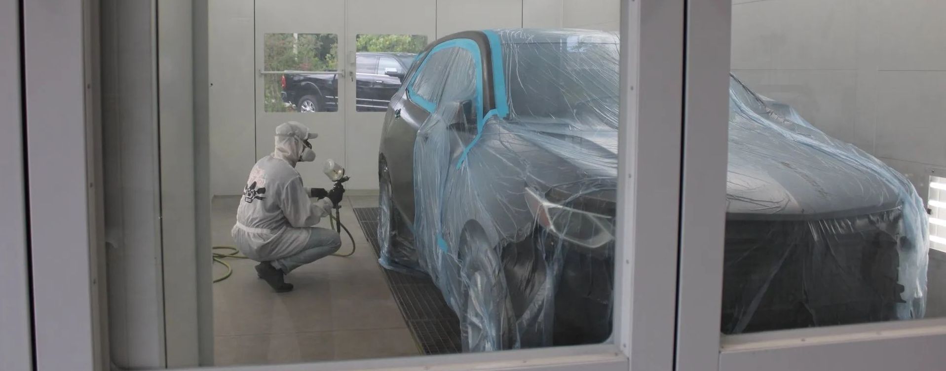 A person in protective gear sprays paint on a car covered in plastic inside a paint booth.