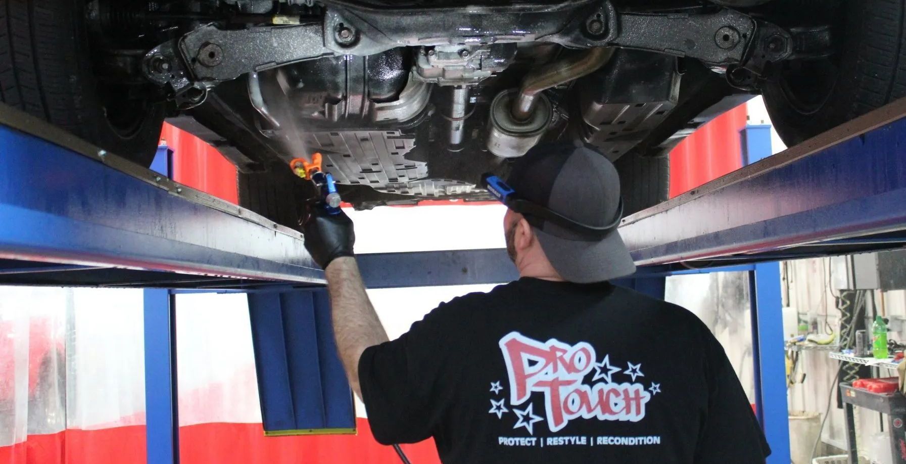 A person sprays rustproofing on the undercarriage of a car raised on a lift in a shop.