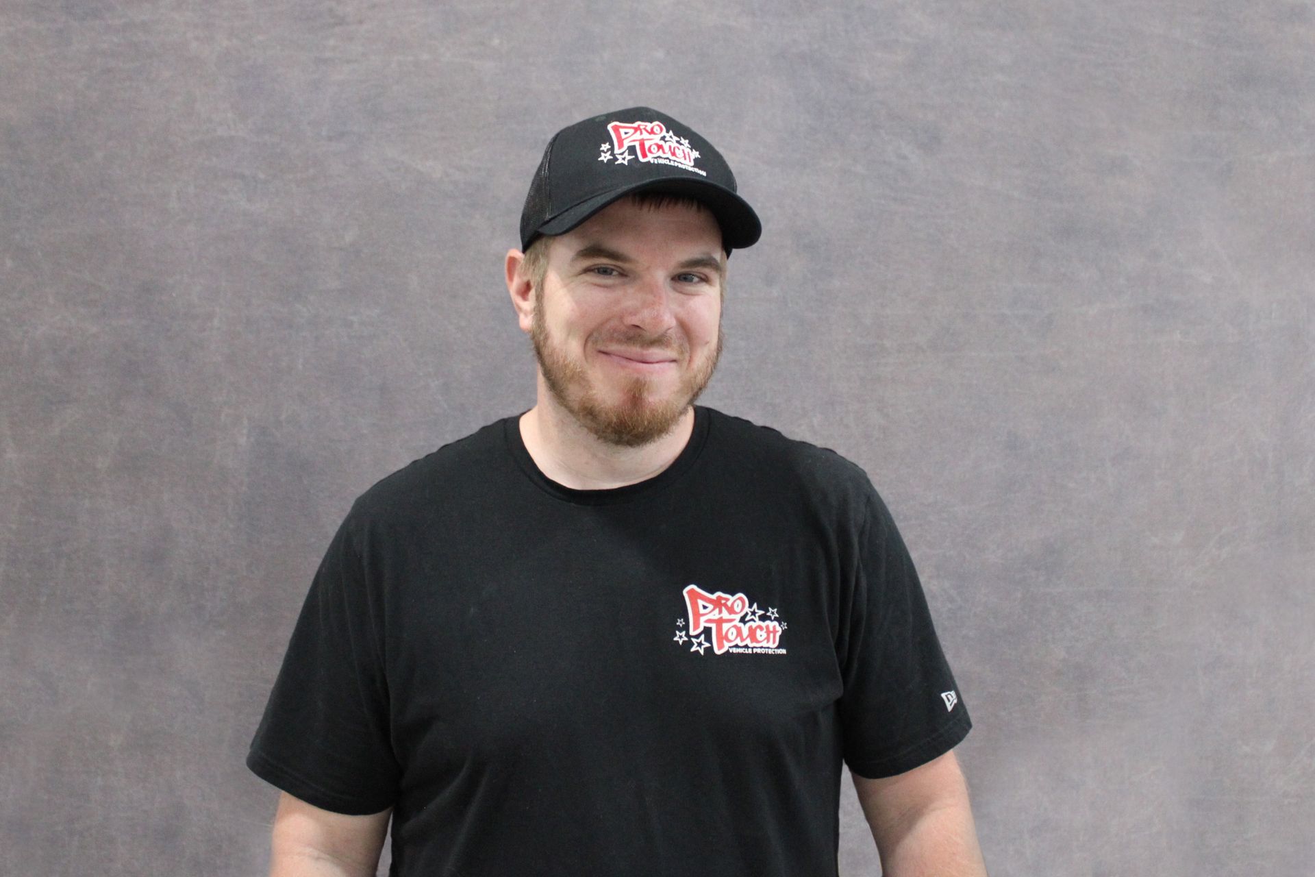 Man in a cap and black hoodie smiles at the camera against a gray background.