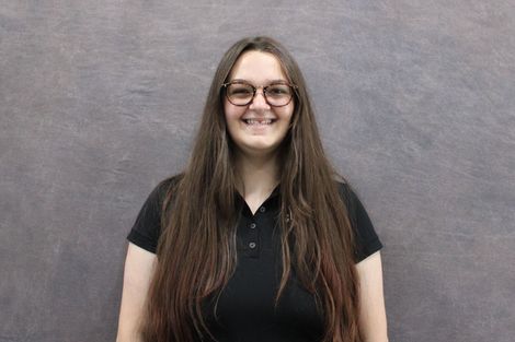 Woman with glasses smiles wearing a black shirt with logo, braids, standing in front of a gray backdrop.