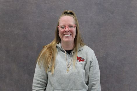 Woman with glasses smiles wearing a black shirt with logo, braids, standing in front of a gray backdrop.