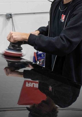 Person polishing a black car with a rotary buffer, indoors.