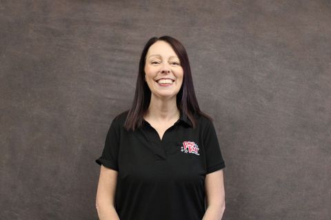 Woman with purple-streaked hair, smiling, wearing a black shirt with a logo, standing against a gray backdrop.