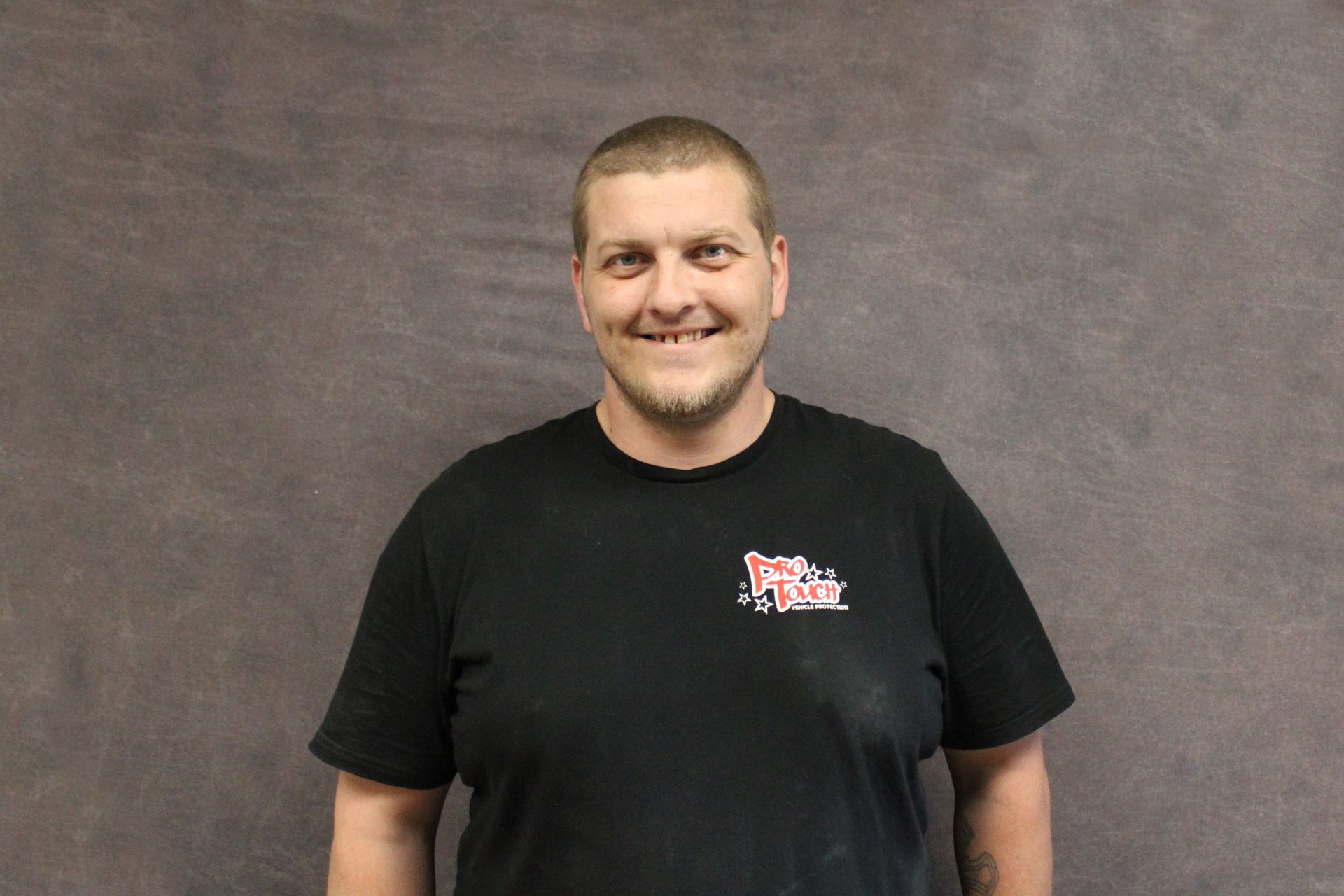 Man in black t-shirt smiles at the camera, standing in front of a gray background.