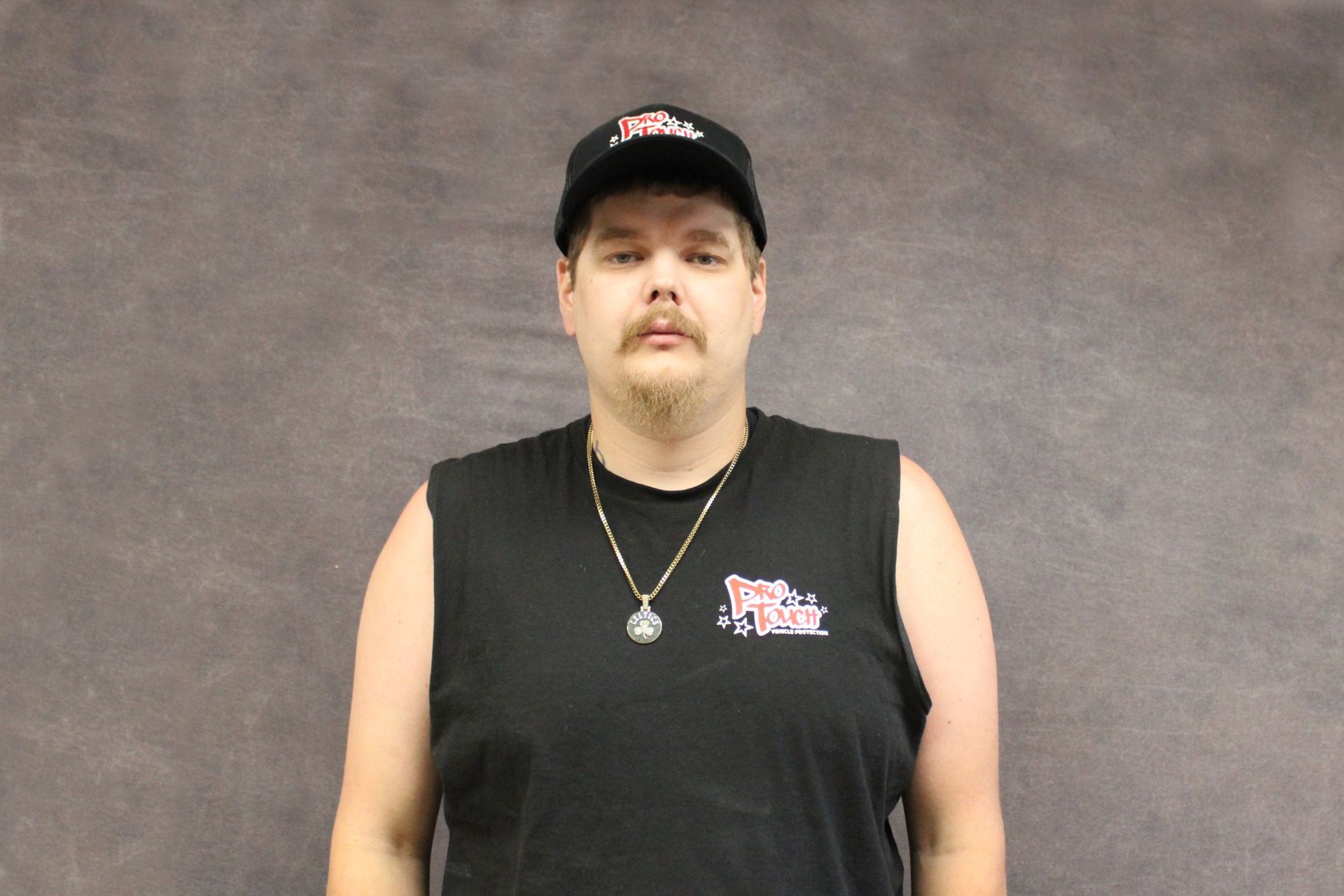 Man with curly hair and mustache in a black t-shirt, wearing a necklace, standing in front of a gray backdrop.