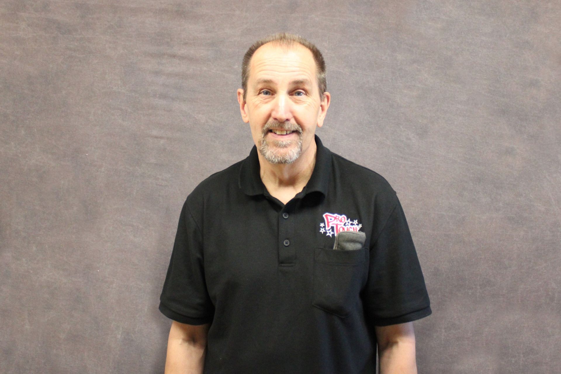 Man in black t-shirt smiles at the camera, standing in front of a gray background.