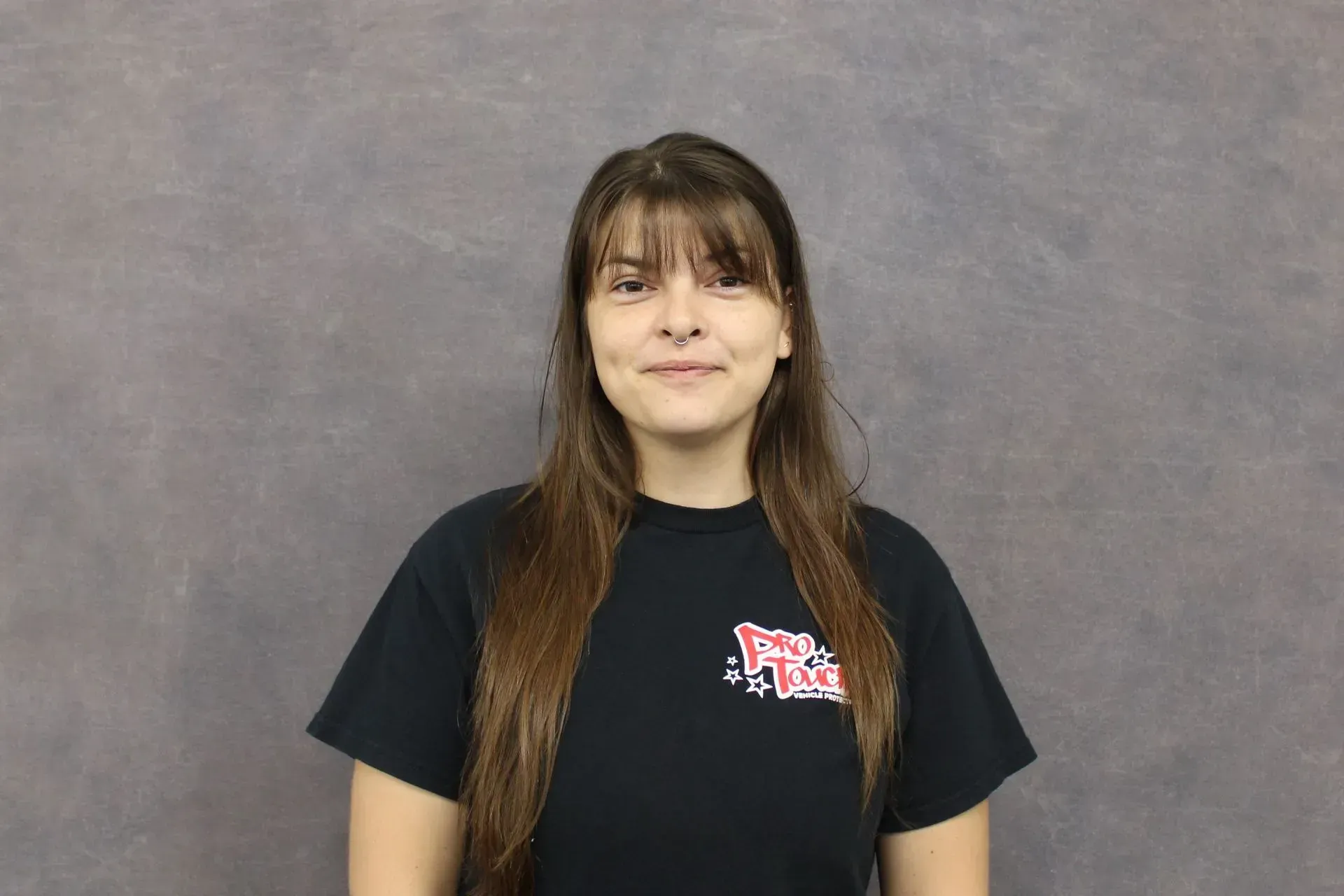 Woman with brown hair and bangs smiles, wearing a black t-shirt with a red logo, standing in front of a gray backdrop.