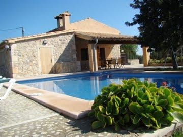 Stone house with a pool, a covered patio with chairs, and lush greenery under a blue sky.