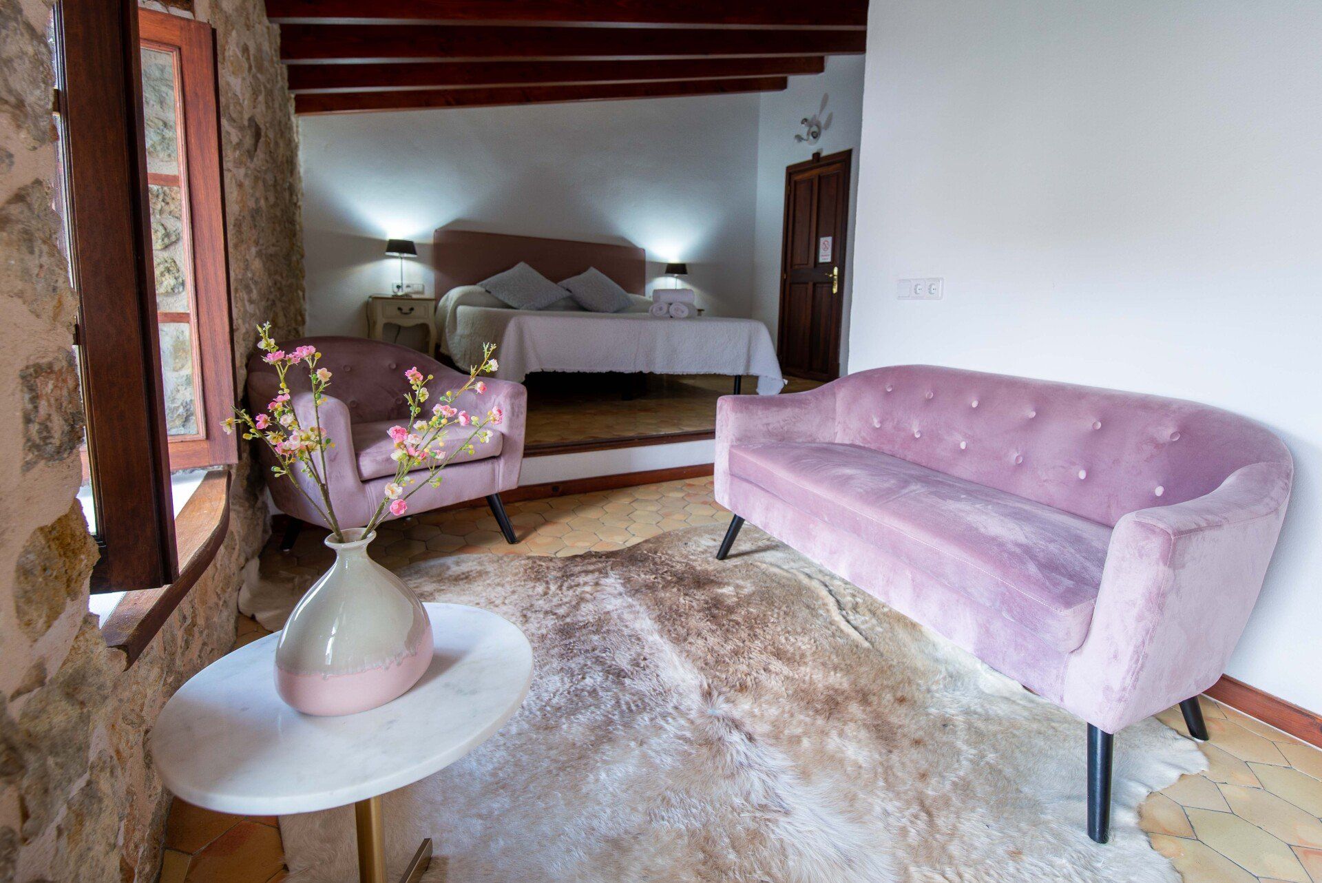 Cozy bedroom with pink sofa and armchair, cowhide rug, stone wall, and white bed.
