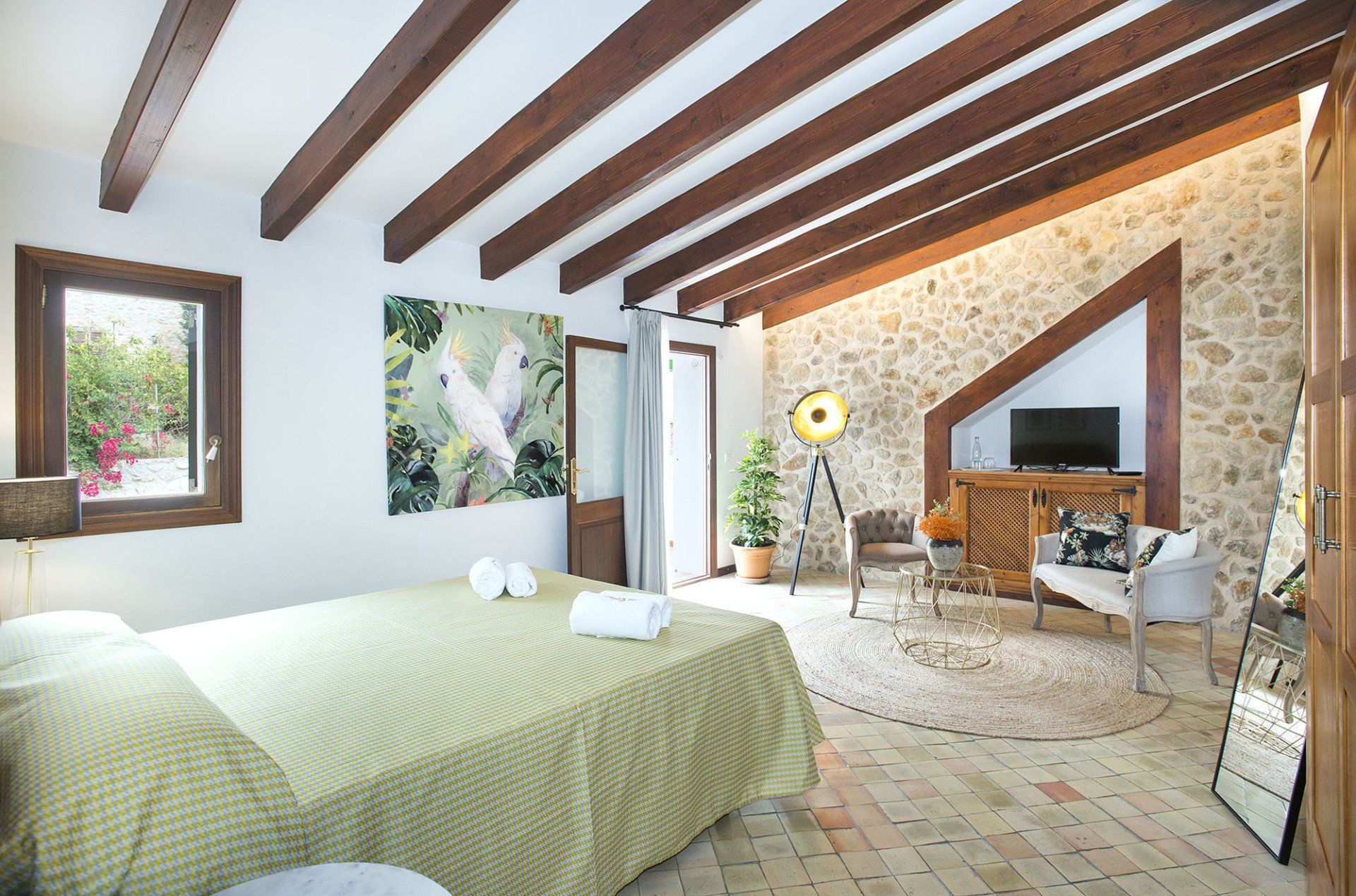 Bedroom with stone wall, wooden beams, bed with yellow cover, art, and seating area.