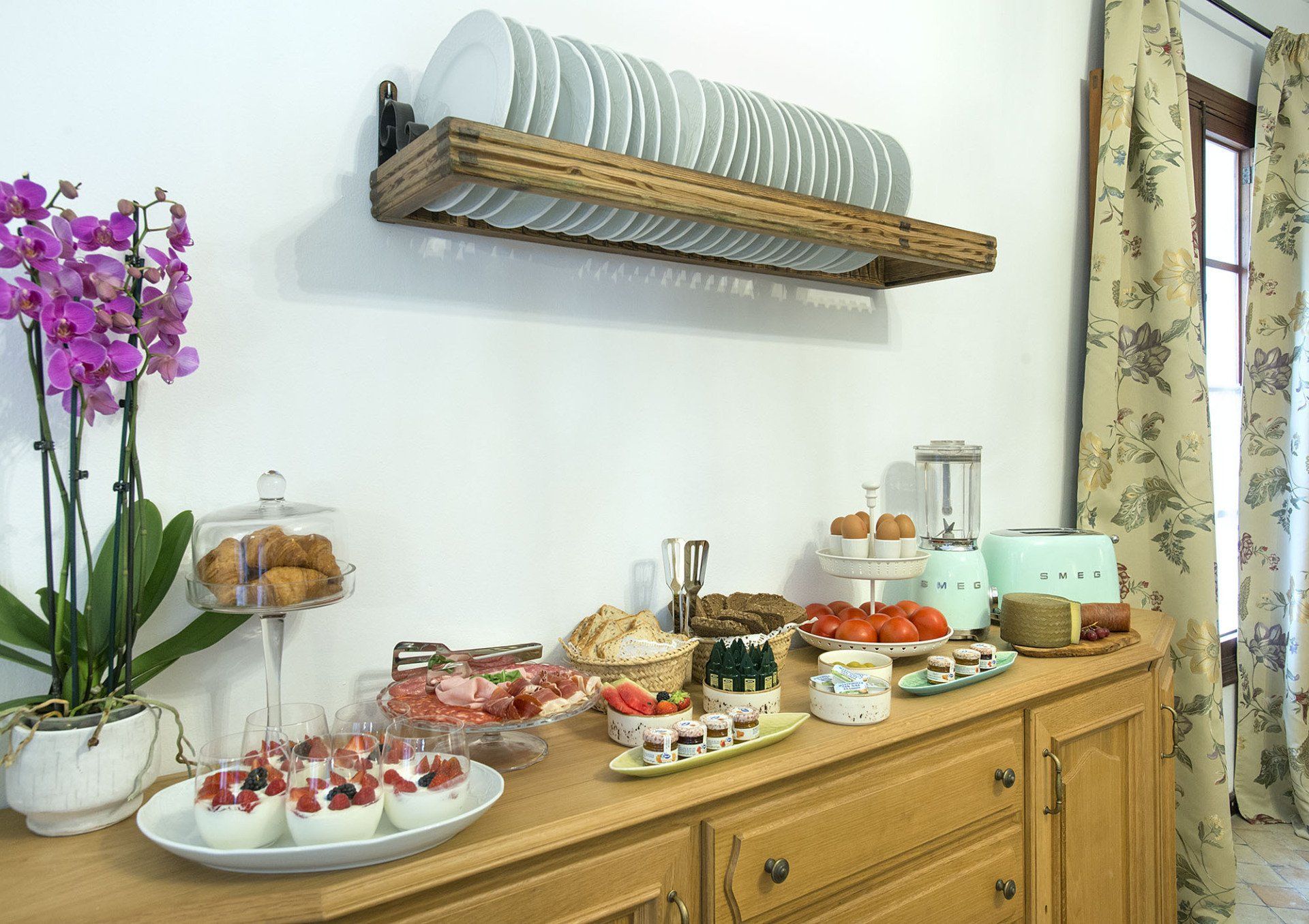 Wooden sideboard buffet with food, plates, and an orchid in front of a window.