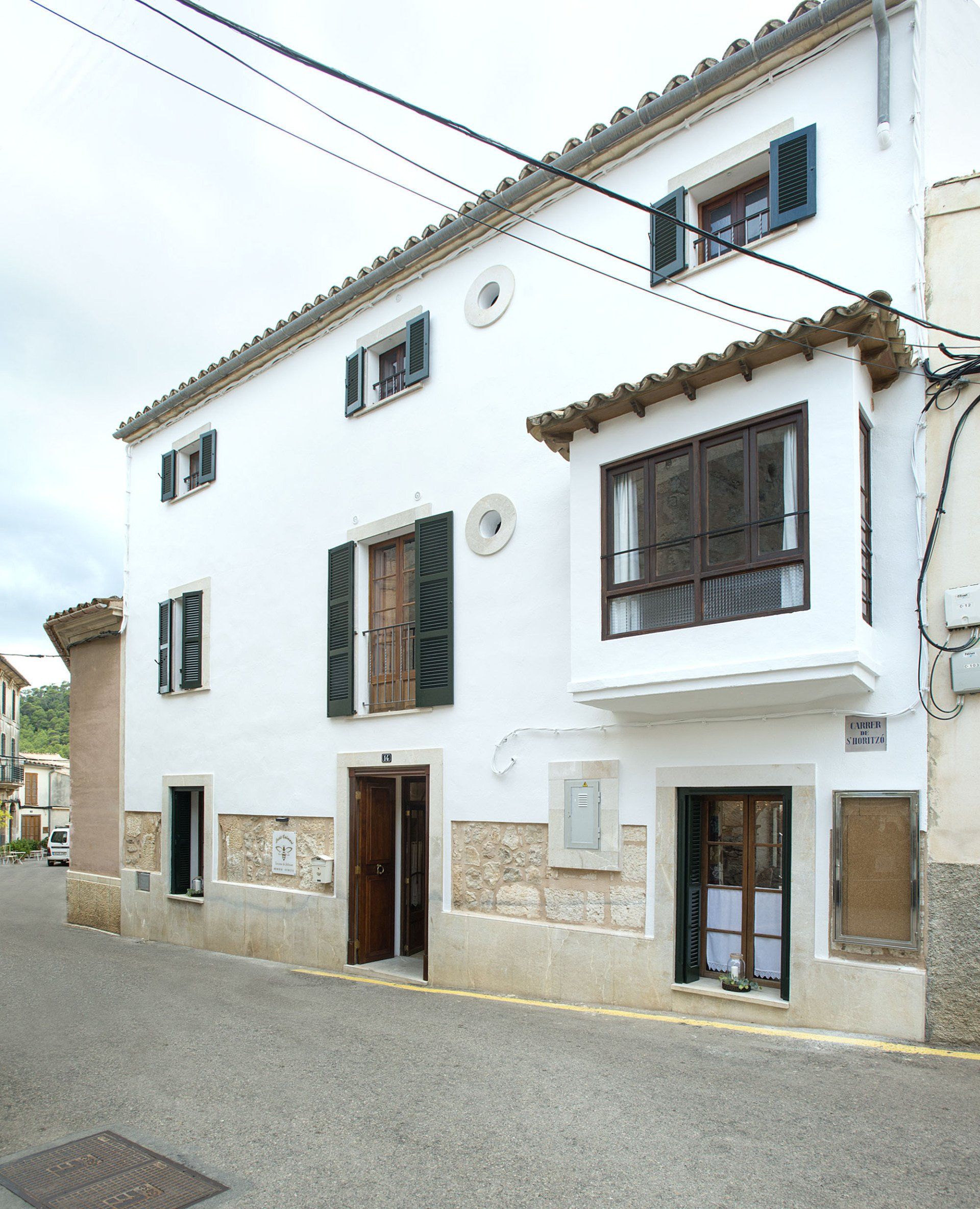 White stucco house on a street, featuring dark window shutters and a small balcony.
