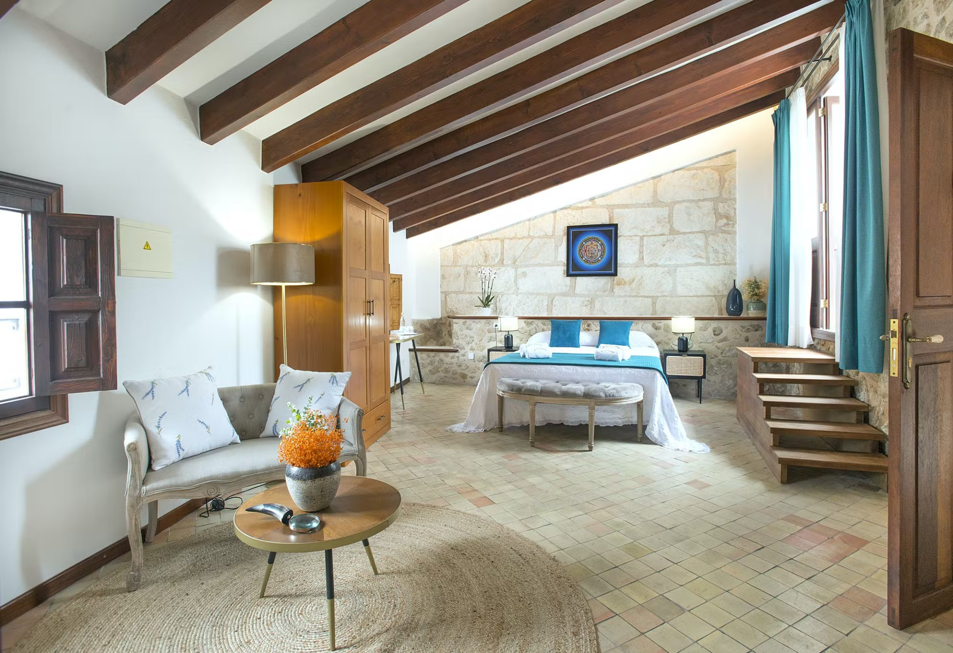 Bedroom with wood beams, stone wall, bed, sofa, and small stairs to a raised platform.
