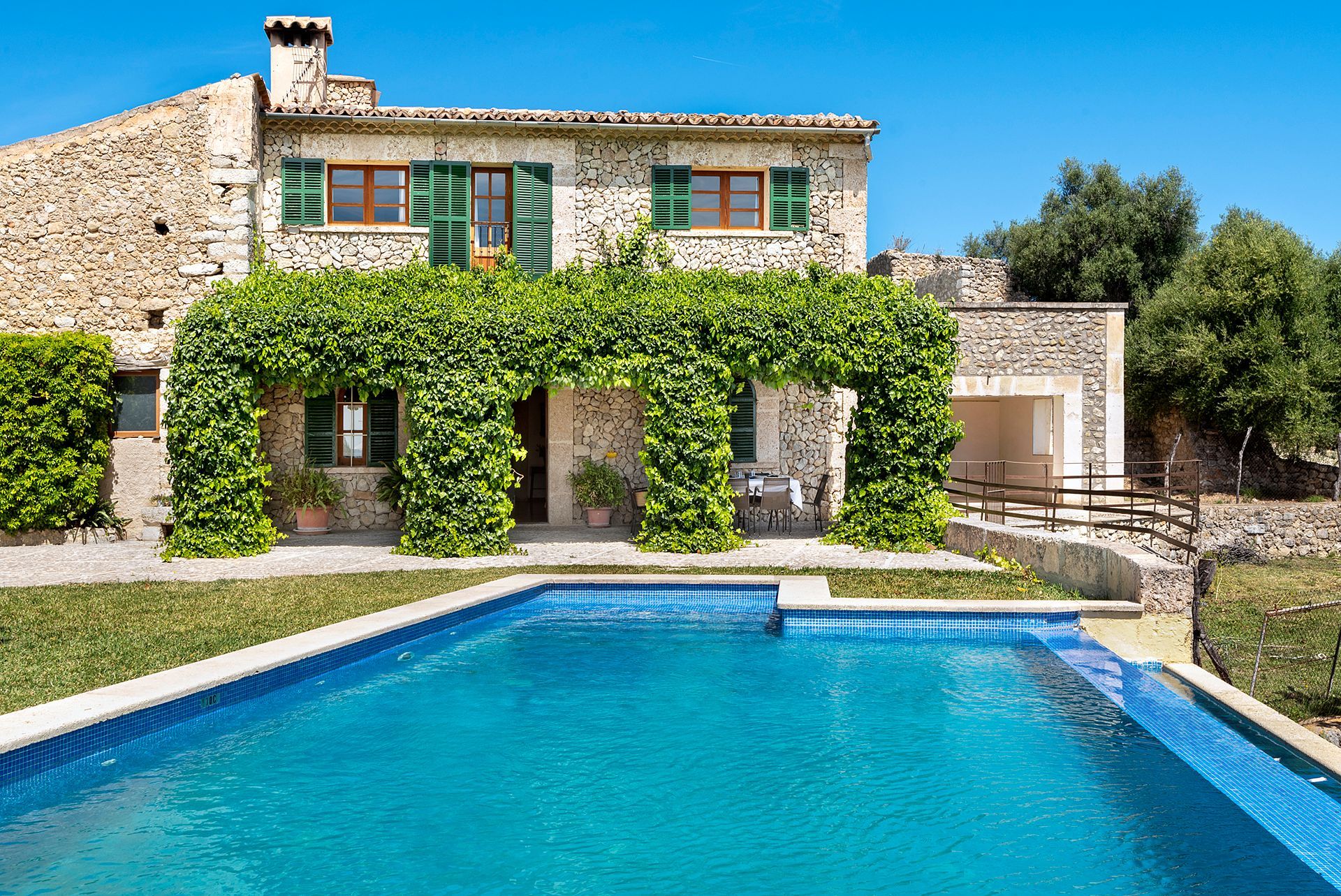 Stone house with a pool, surrounded by greenery and a blue sky.