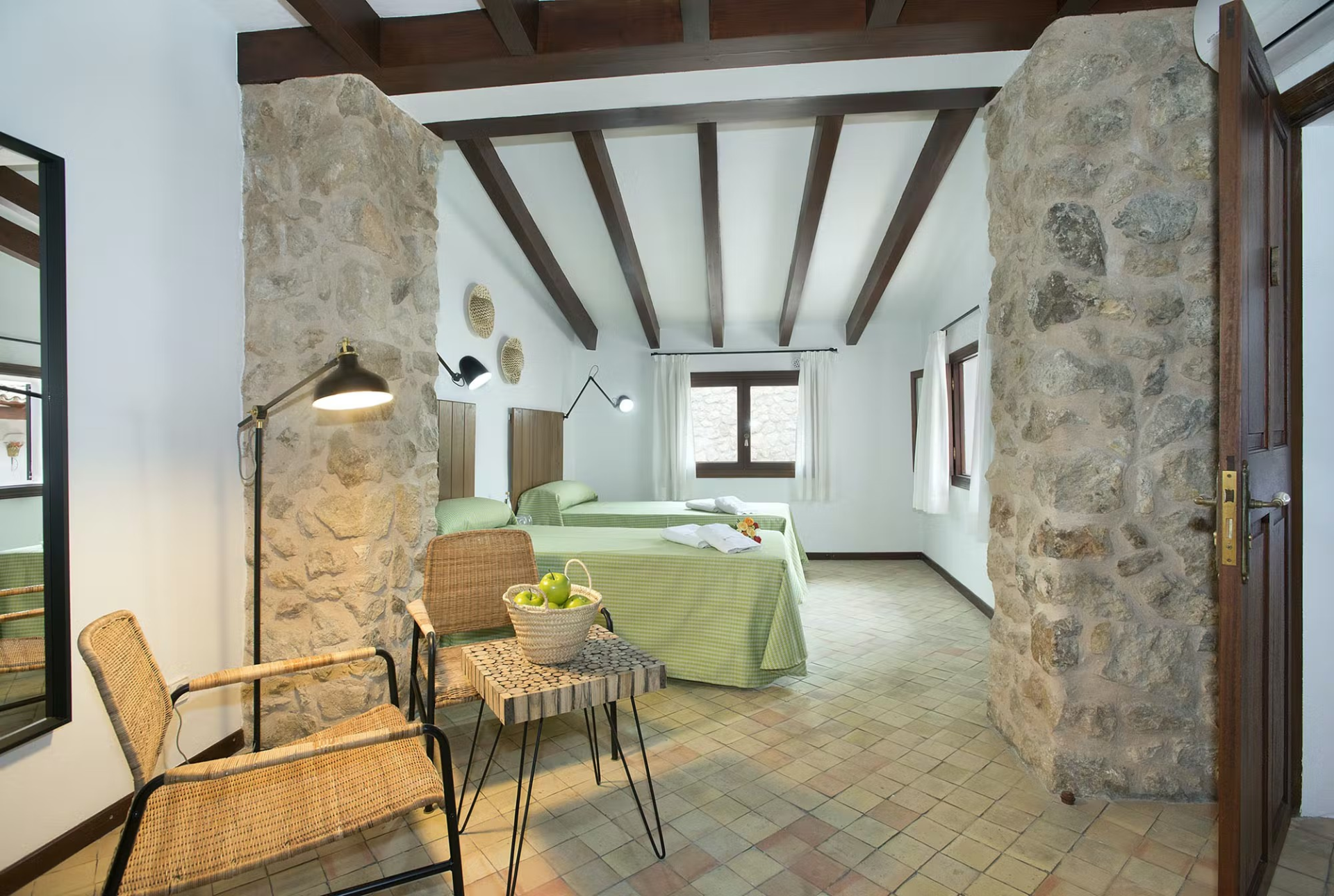 Bedroom with two beds, stone columns, wooden beams, wicker chair, and small table with fruit.