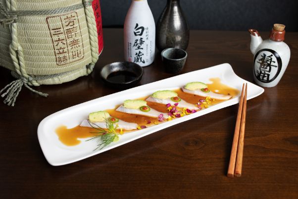 A long white plate of fish slices (Hamachi Carpaccio) with chopsticks on a wooden table.