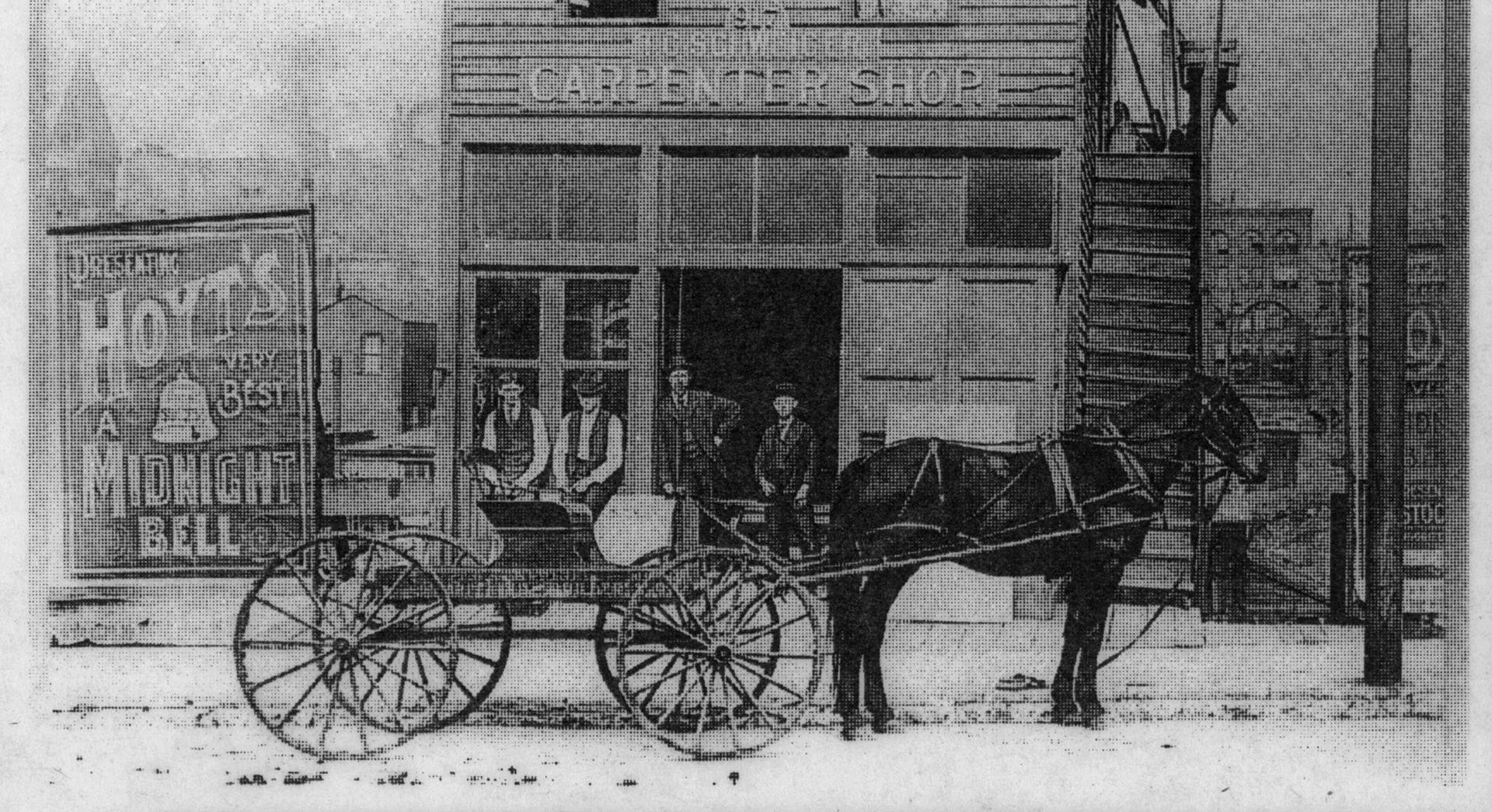 A black and white photo of a horse drawn carriage in front of a building.