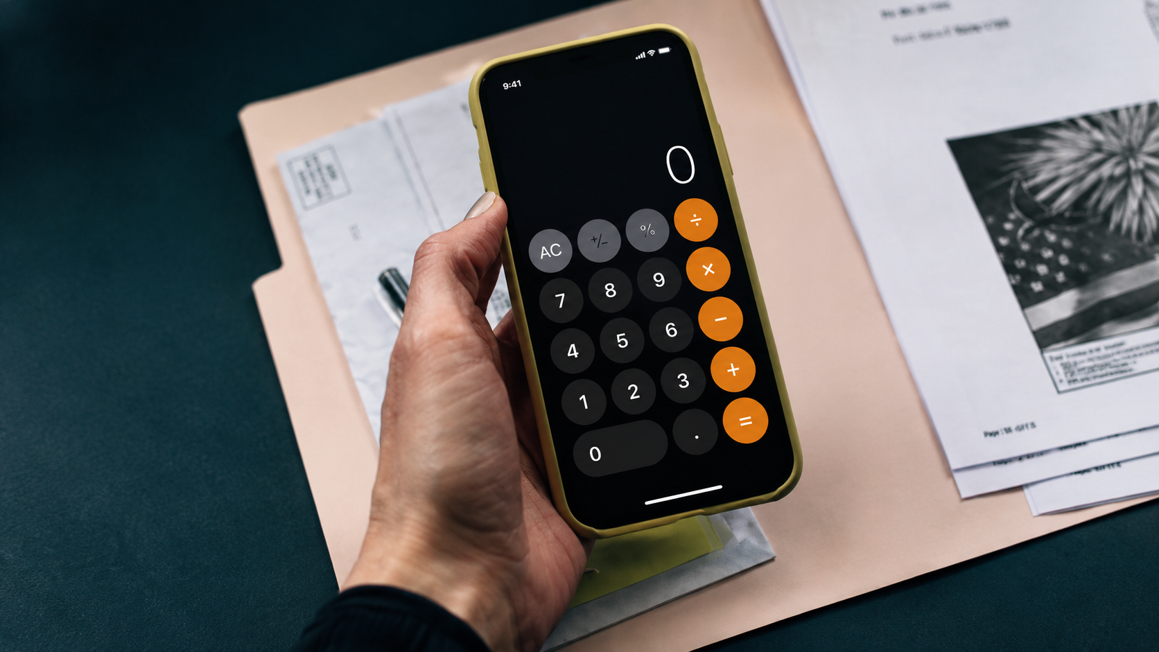 Hand holding a smartphone with a calculator app over papers on a desk